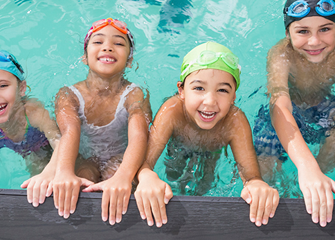 Smiling kids in swim caps and goggles lined up in the water along the edge of a pool.